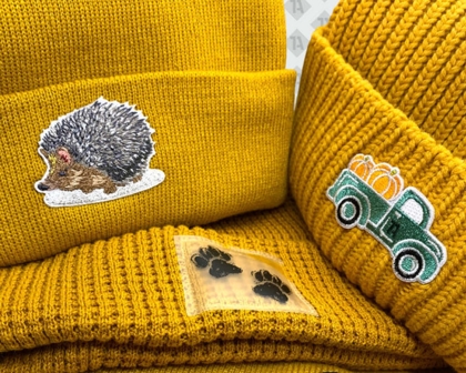 Drei senfgelbe Beanies mit gestickten Patches: Igel, Truck mit Kürbissen und Pfotenabdruck.