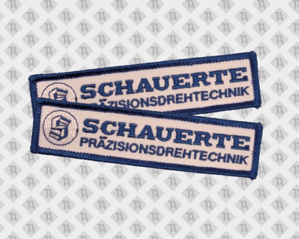 Weiß-blauer Namensaufnäher mit Schauerte-Logo und der Aufschrift Präzisionsdrehtechnik, dargestellt in zwei Varianten