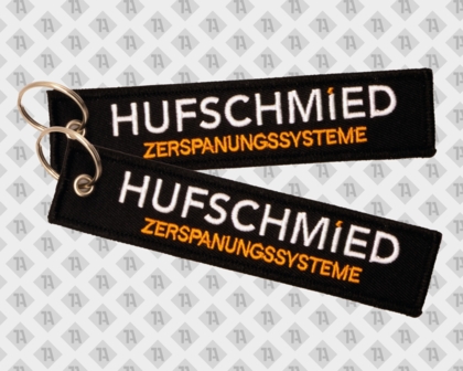 Schwarzer HUFSCHMIED ZERSPANUNGSSYSTEME Schlüsselanhänger, rechteckiger Stofftag mit weißer und orangefarbener Stickschrift und Metallring