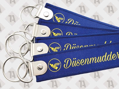 1A Schlüsselanhänger in Blau, gewebt mit gelber Schrift, Kettelrand und Metallring.