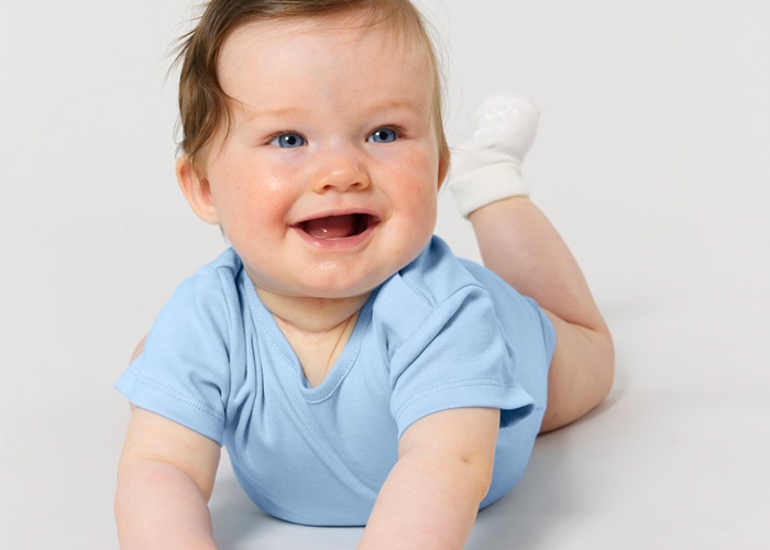 Baby in himmelblauem Kurzarm-Body liegt auf dem Bauch und lacht.
