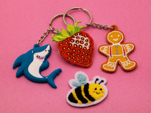 Einige Beispielartikel aus dem 1A-Shop auf rosafarbenen Untergrund: Schlüsselanhänger Hai und Lebkuchenmann, ein Erdbeer-Pin und eine Chenille-Hummel