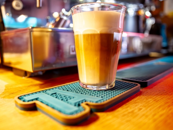 Unser 1A-Gummi-Untersetzer mit 1A-Logo in auf einem Tresen mit einem Glas Milchkaffee