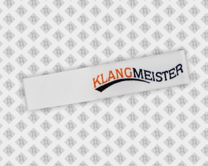 Gewebtes Label mit Laserschnitt – KLANGMEISTER in Schwarz und Orange auf Weiß