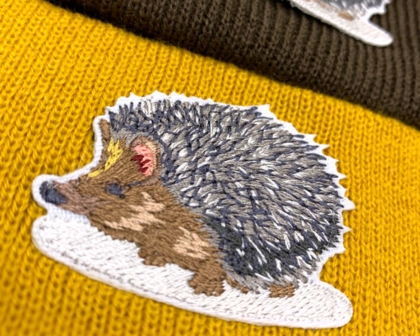 Gestickter Igel-Patch auf einer gelb-braunen Beanie, detaillierte Stickerei mit Naturmotiv.