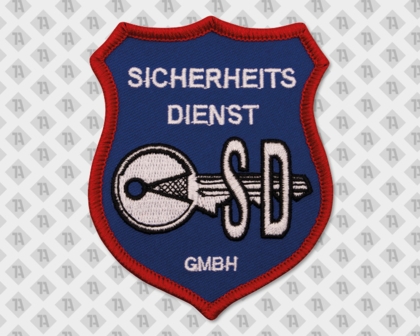 Blauer Schild-Patch mit rotem Rand, weißem Schriftzug „Sicherheitsdienst GmbH“ und einem großen gestickten Schlüssel mit „SD“.